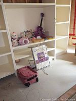 Image of white shelving with various items including the wood dollhouse kit box, pink backpack on floor, purple ukulele, Hello Kitty CD player, stuffed animal, and miscellaneous items.