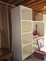 Front view of the tall white shelving unit consisting of two attached parts with multiple open shelves, placed in an unfinished basement area.