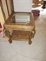 Side view of ornate carved wood side table with glass top and lower shelf, showing gold-tone finish and claw feet.