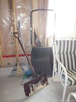 Photo showing 8 garden tools including a large metal scraper, metal rake, plastic snow shovel, broom and rakes stored in basement.