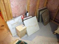 Tiles of various sizes are stacked against an unfinished wall in basement, showing several boxes and individual tiles, some with marble-like veining.