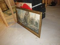 Side view of large ornate framed print of forested area with lake and mountains, measuring 30x54 inches. Behind it, the corner of a square stretched canvas print of a red flower, approx 38x38 inches.