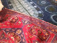 Photo shows portion of large red decorative rug with octagonal and diamond shaped floral and geometric patterns and fringe on one edge, placed partially over a blue rug with circular patterns.