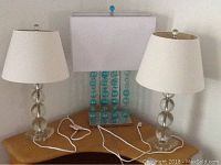 Set of three lamps on wooden surface against white wall, showing two clear sphere-based lamps with white shades and one blue decorative lamp with bead base and rectangular shade