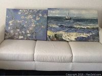 Two oil paintings placed side by side on a white couch: one of white blossoms on tree branches, the other of a sailboat on the sea.