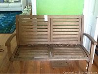 Front view of wood bench showing slatted backrest and seat with armrests, sitting on hardwood floor indoors.