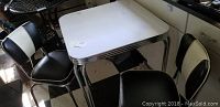 Photo showing 1950s style square table with light-colored top and chrome edges with two retro-style chairs with black and white padded seats and backs.