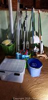 Photo showing assortment of cleaning supplies including brooms, brush, various mops, green basket with items, blue bucket with wringer, and transparent storage box