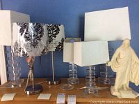 Four acrylic table lamps with stacked square block bases and white fabric shades, two chrome table lamps with patterned shades, and a cream resin statue on a shelf against blue wall.