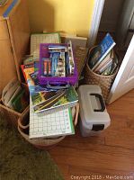 Overview of lot showing baskets filled with art books and supplies, plastic toolbox, and easel box leaning against wall