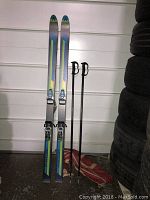 Pair of Dynastar Mystere skis leaning against wall, accompanied by two Scott ski poles and a red-beige ski bag on the floor.
