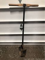 Full view of the post hole digger showing wooden handles, black metal shaft, and digging blades with rust.