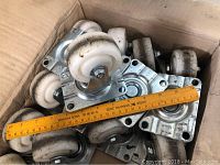Box containing twelve white nylon swivel wheel castors with metal mounting plates and ruler showing size.