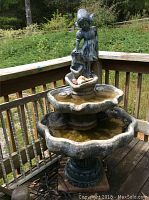 4-tier concrete patio fountain with girl statue pouring water at top, shown on wooden deck with surrounding yard.