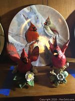 Three cardinal bird figurines and one decorative plate in background, showing detailed red and natural cardinal colors, handcrafted details, and floral bases.