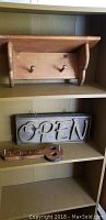 Photo showing small wooden wall shelf with two pegs, wooden 'OPEN' sign, and small stringed instrument shaped decorative piece on shelf
