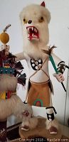 Tall Native American doll in white fur with painted facial features holding feathers and a stick.