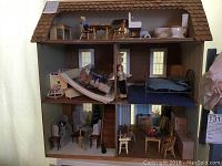Front view of three-story wooden doll house with various rooms furnished including living room, bedroom, and dining area. Two dolls visible inside.