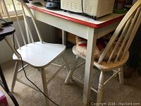 Photo of the vintage kitchen table with porcelain top and red trim showing the table with two white wooden chairs and part of a countertop appliance.