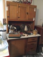 Front view of modified Hoosier cabinet showing porcelain top with various kitchen items on it, upper three-door wooden cabinet, and lower cabinet with drawers