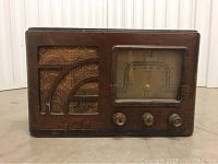 Front view of antique Stomberg Carlson radio receiver with wooden case, fabric speaker cover, and analog dial with three knobs beneath.