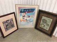 Three framed artworks standing on floor against wall: Tahiti map, Corn Hill Arts Festival poster, and angel with deer print.