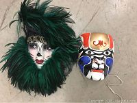 Two full-face painted masks; one with green feathers and black lace, the other painted in orange, black, blue, and white aping a tribal or theatrical style.