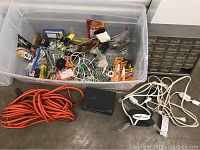 Photo showing a large clear plastic storage bin containing multiple small hardware packages, fasteners, and small tools, with several tangled white electrical cords and one orange extension cord lying outside the bin on the floor.