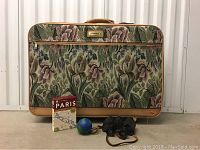 Photo showing the full rolling carpet bag with floral tapestry, Paris City Walks guide, stress ball, and vintage binoculars in front.