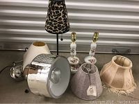 View of all lamps and lampshades showing two white ceramic lamps, silver reflector lamp, beige and gray lampshades, and leopard print shade on stand