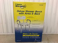 Photo of unopened box showing product details for Essential Bath Safety Deluxe Shower Bench with Arms & Back model 8301, highlighting features like curved back, padded arms, and non-slip seat.