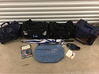 All 5 bags lined up with accessory items on floor in front: 4 duffel/carry-on bags in blue and black, Victoria's Secret tote, umbrella, and EDS logo item.