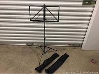 Photo showing one black metal music stand fully assembled, with tripod legs extended and music plate in place, alongside two black travel/storage bags for the stands.