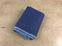 Photo showing a folded blue padded furniture moving blanket with quilted zigzag stitch pattern and contrasting light blue binding edges lying on a concrete floor.