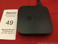 Top view of black Apple TV device with visible Apple logo and 'tv' marking on top, next to MaxSold auction ticket