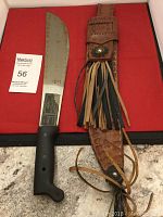 Knife with black handle and large blade alongside tassel leather sheath with button closure on red background.