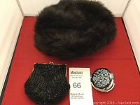 Front view of dark brown faux fur hat, black beaded purse with gold-tone clasp, and compact mirror with black floral lid on red surface.