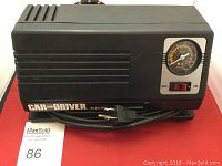 Black Car and Driver electric air compressor with pressure gauge and on/off switch visible. Power cord coiled underneath, showing standard two-prong plug.