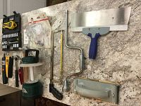 Top view showing all tools laid out on a stone surface including hand crank drill, scrapers, tape measure, hacksaw frame, utility knife, and camping lantern