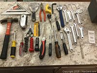 Variety of hand tools laid out on a countertop including hammer, screwdrivers, pliers, wrenches, hex keys, scissors, socket bits, and a carpenter's square.