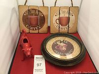Two coffee-themed square wall hangings with coffee cup images and text, a round coffee-themed clock, and a red ceramic cow creamer holder on a red surface