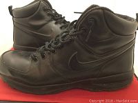 Side angle of the pair of black Nike Manoa sneakers showing profile and soles, placed on a red surface.