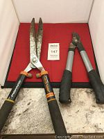 Fiskars hedge clipper and pruning shears placed side by side on red surface showing overall condition and branding.