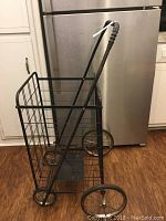 Black metal rolling hand cart with wire basket and rubber handle grip against kitchen background.