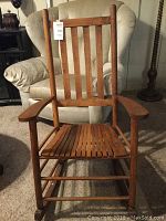 Front view of wooden rocking chair showing slatted seat and vertical slatted backrest, placed on carpet with beige upholstered armchair in background.