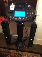 Full view of the karaoke system showing main machine with base, two side speakers, digital display illuminated in blue, keypad, and control knobs.