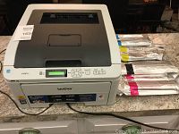 Front and side view of the Brother HL-3075CW color laser printer with four additional toner cartridges in original packaging.