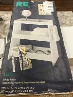 Packaging showing image of assembled white finish bathroom cart with three shelves and towel holder, size 23.5x19x9.25 inches, hardware included, assembly required.