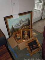 Four paintings leaning against wall and in cardboard box: one large alpine scene and three smaller framed paintings, two showing elderly men with mugs and one showing a group around a table.