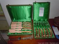 Two open original green textured boxes showing sets of knives and forks with decorated metal and antler horn handles.